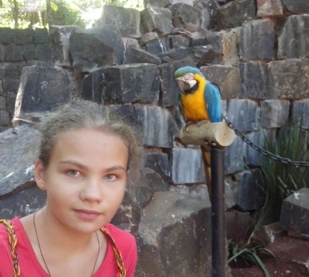 Menina loira ao lado de arara e pedras cinzas ao fundo