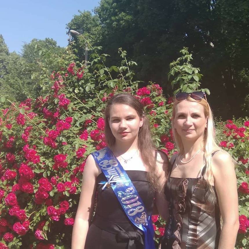 Mulher e menina com faixa azul na roupa em frente a arbusto com flores rosas
