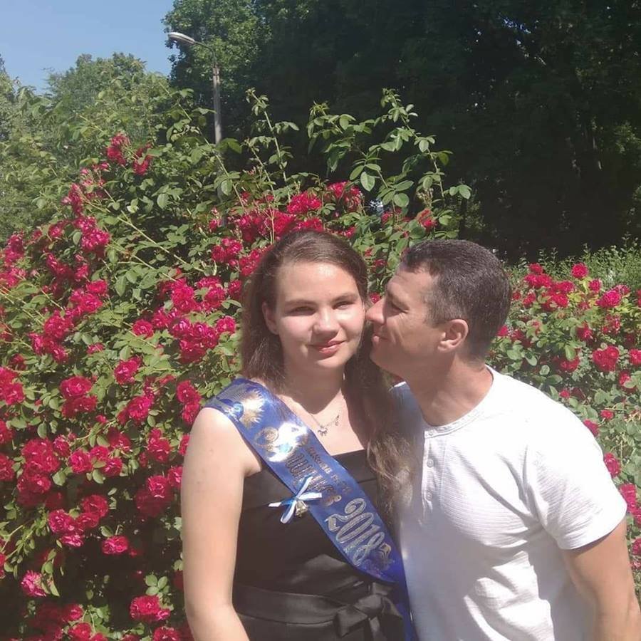 Homem beijando rosto de menina com faixa azul na roupa em frente a arbusto com flores rosas