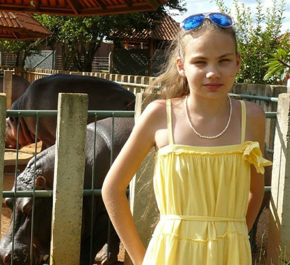 Menina de vestido amarelo e óculos azul na cabeça em frente a hipopótamos em zoológico