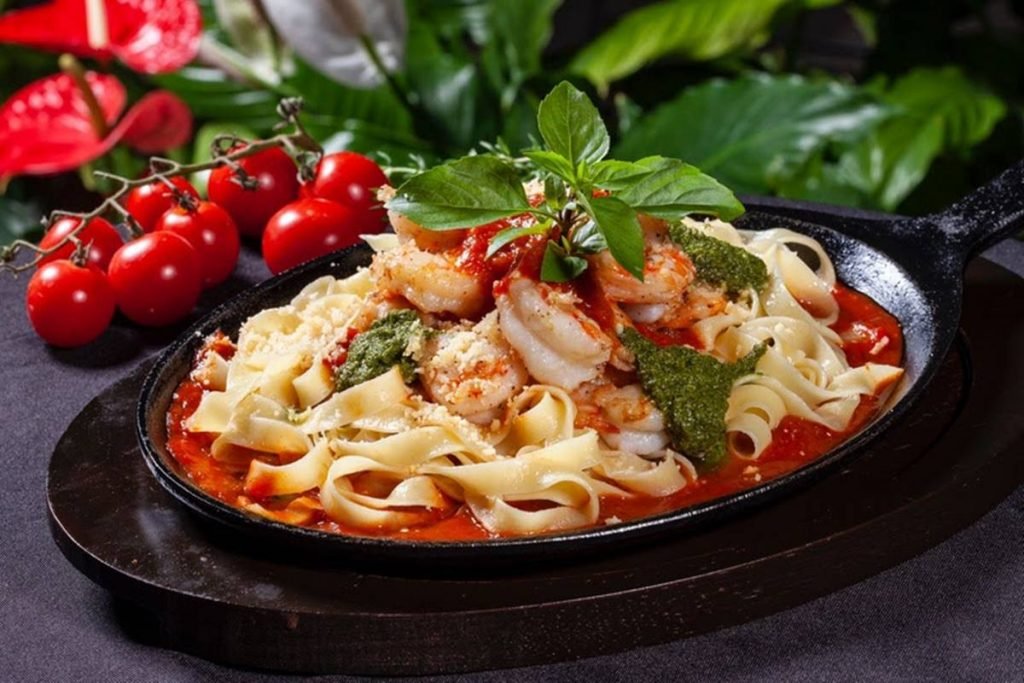foto de prato preto com macarrao, molho vermelho, camaroes e folhas verdes e ao fundo, tomatinhos e folhas verdes