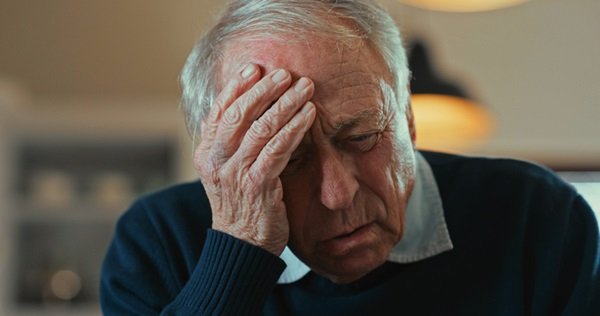 Elderly man with hands on head.  He wears a dark coat, has white hair and looks down- Metropolises