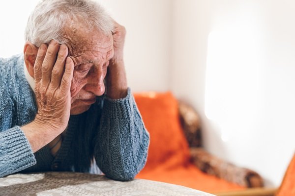 Elderly man with hands on head.  He wears a dark coat, has white hair and looks down- Metropolises