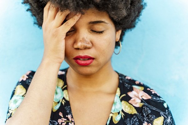 Woman with a hand on her head.  She wears a flowered shirt, golden earrings and red lipstick- Metropolises