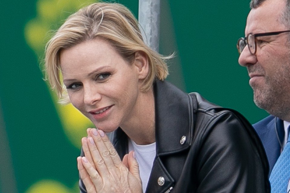 Foto colorida. Princesa Charlene com as mãos juntas e uma jaqueta de couro