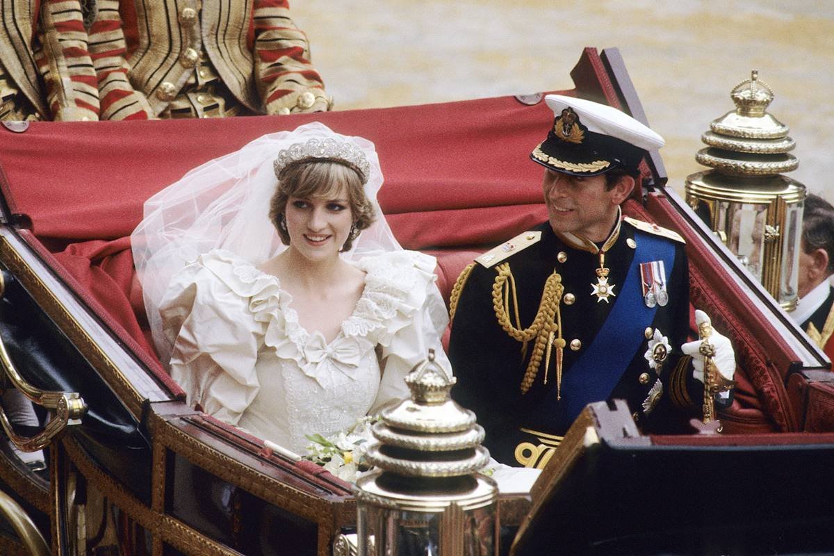 Princesa Diana e príncipe Charles do Reino Unido em carruagem real no dia de seu casamento. A princesa usa vestido e coroa e o príncipe usa farda militar - Metrópoles
