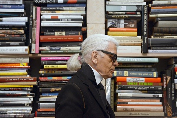 Karl Lagerfeld, estilista da Chanel, em sua biblioteca. Ao fundo, estantes com livros. Ele usa blazer preto, camisa branca e óculos escuros