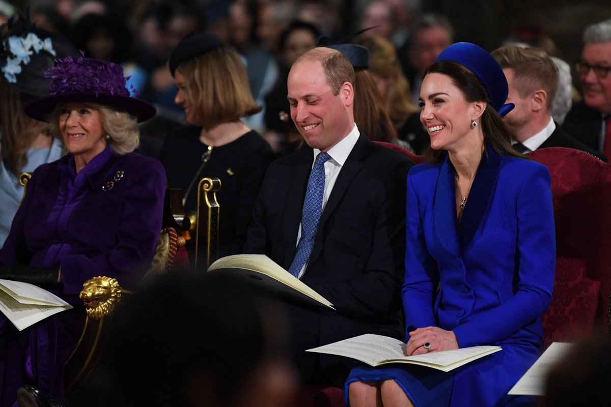 Foto colorida. Camilla Parker, príncipe William e Kate Middleton estão sentados e seguram papéis