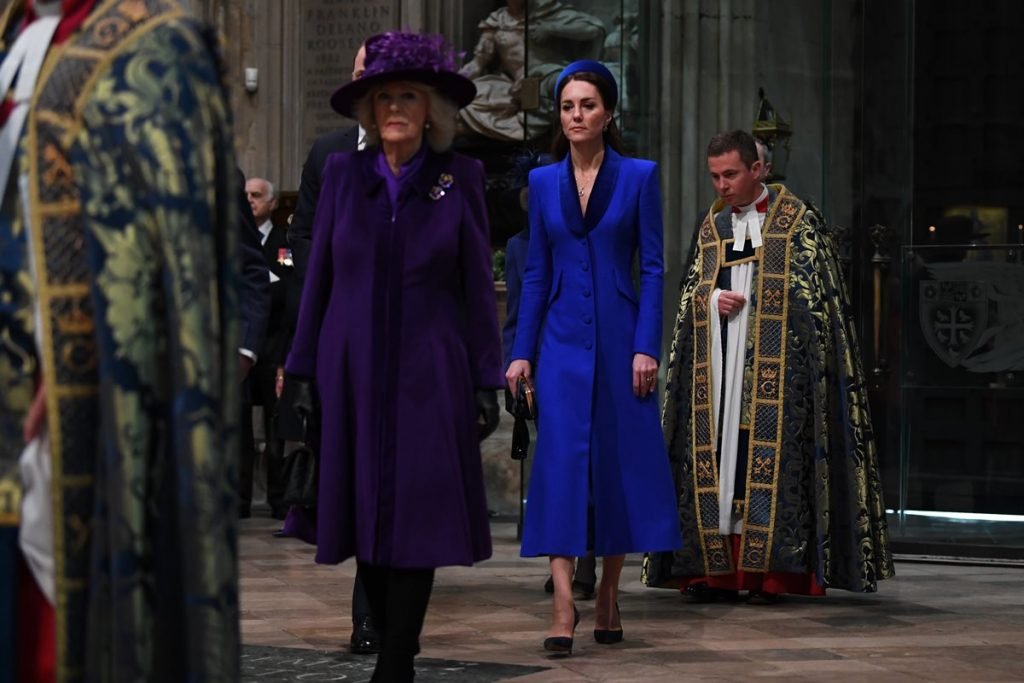 Foto colorida. Camilla Parker e Kate Middleton. A primeira veste um casaco roxo e a segunda, azul