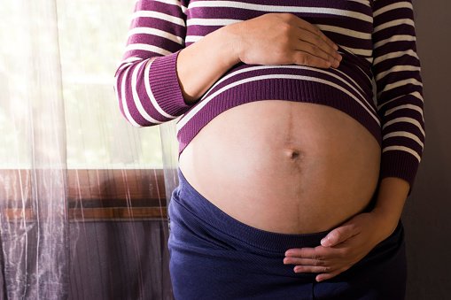 Mulher grávida mostrando a barriga com uma listra no meio em frente à uma janela