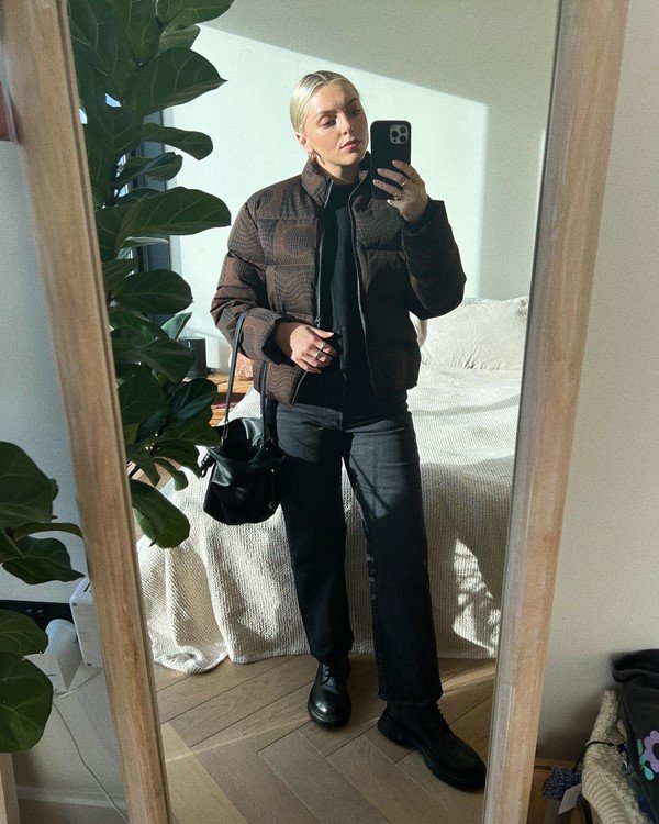 Mulher branca, com cabelo louro amarrado, tirando foto com celular em frente ao espelho. Ela usa calça, botas e blusa pretas, e um casaco marrom.
