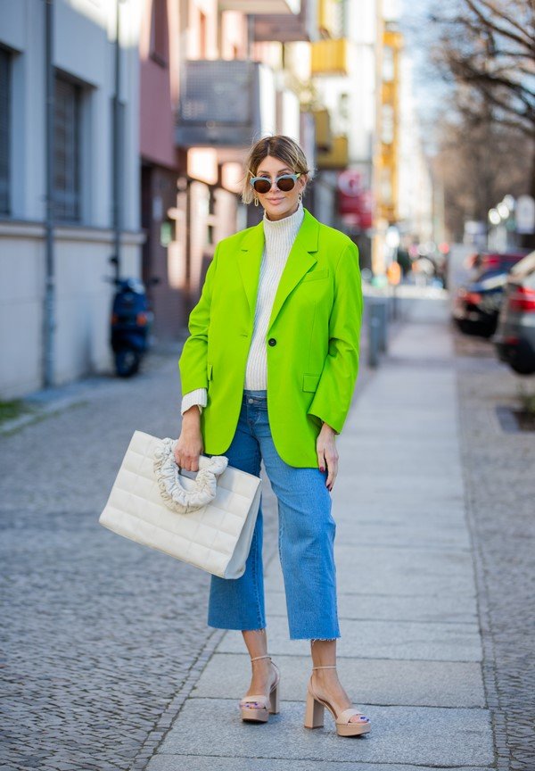 Mulher branca e loura com cabelo amarrado posando na rua. Ela usa uma calça jeans, um casaco e uma bolsa brancos e um blazer verde limão.