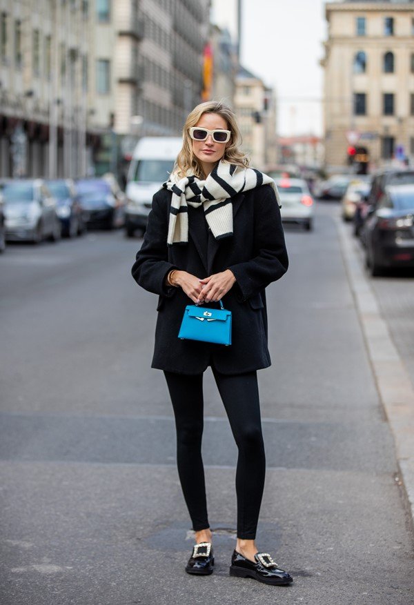 Mulher branca e loura posando na rua. Ela usa calça jeans justa e casaco pretos, um cachecol preto e branco e uma bolsa mini na cor azul.