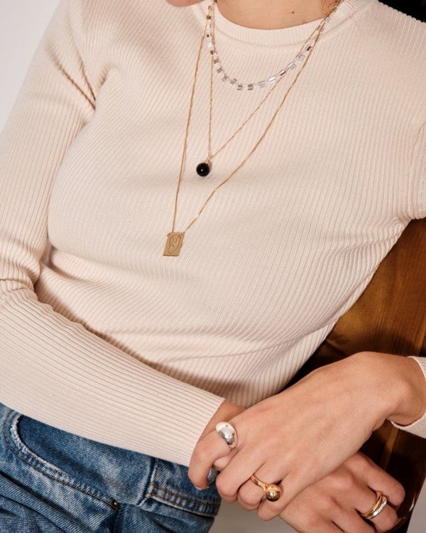 Foto de campanha da marca de acessórios Luiza Dias 111. A mulher branca usa blusa de manga bege, calça jeans e colares e anéis dourados e pratas.