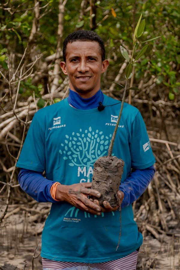 homem negro usa camisa azul, sorri e segura uma muda de planta