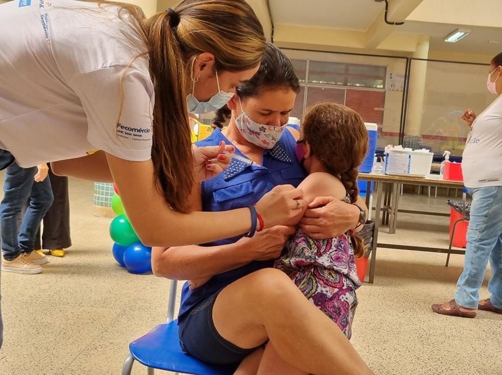 Criança sentada em colo de mulher, com a cabeça para trás mulher enquanto aplica vacina em seu braço