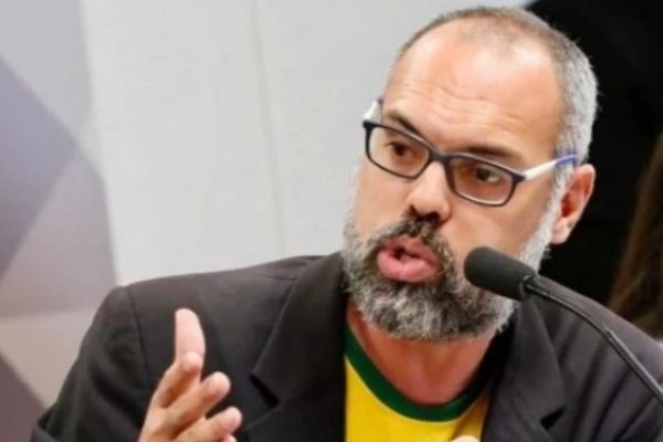 O blogueiro bolsonarista Allan dos Santos fala em Comissão das Fake News, dando seu depoimento sobre acusações de disseminação de notícias falsas. Ele fala em microfone e usa camisa verde e amarela sob paletó - Metrópoles