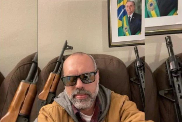 Allan dos Santos, ativista bolsonarista, tira foto ao lado de armas em seu sofá. Atrás, na parede, uma foto do presidente Bolsonaro - Metrópoles