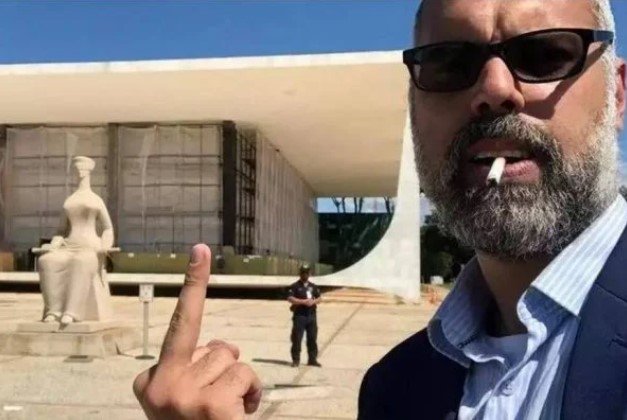 Allan dos Santos, ativista bolsonarista, dá o dedo para o Supremo Tribunal Federal em frente ao prédio.  Ele fuma e usa terno - Metrópoles
