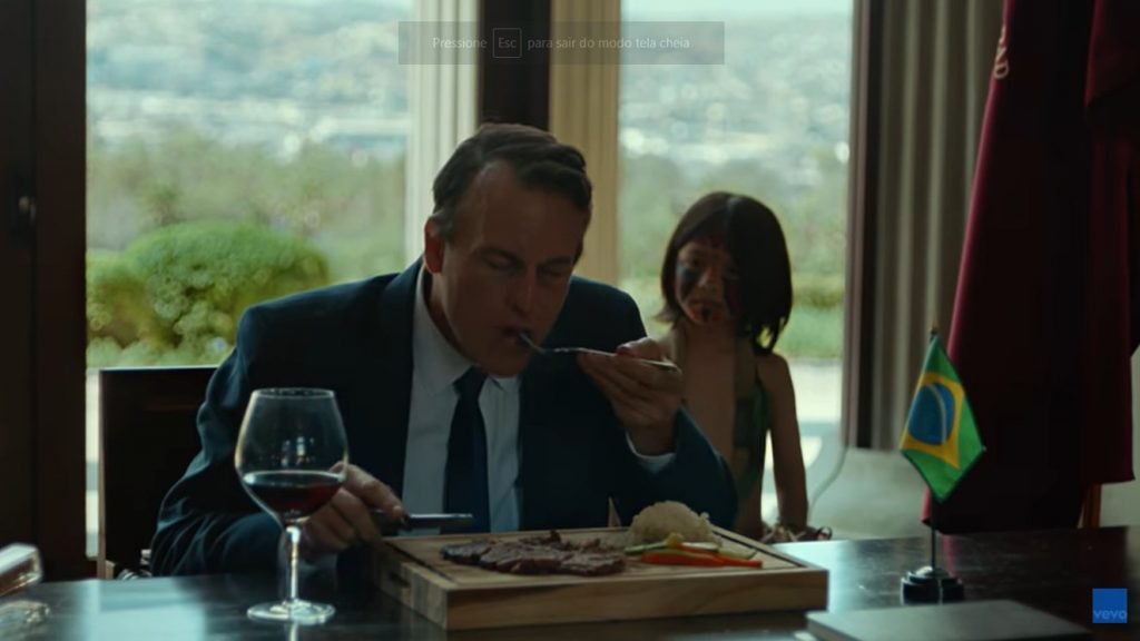 Homem branco de terno come refeição em sala luxuosa, vestindo terno, com uma bandeirado Brasil sob a mesa. Atrás, uma menina indígena olha - Metrópoles