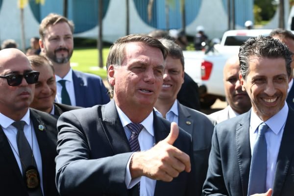 Presidente Bolsonaro na saída do Planalto conversa com jornalistas, fazendo gesto com a mão. Ele está cercado por seguranças e ministros - Metrópoles