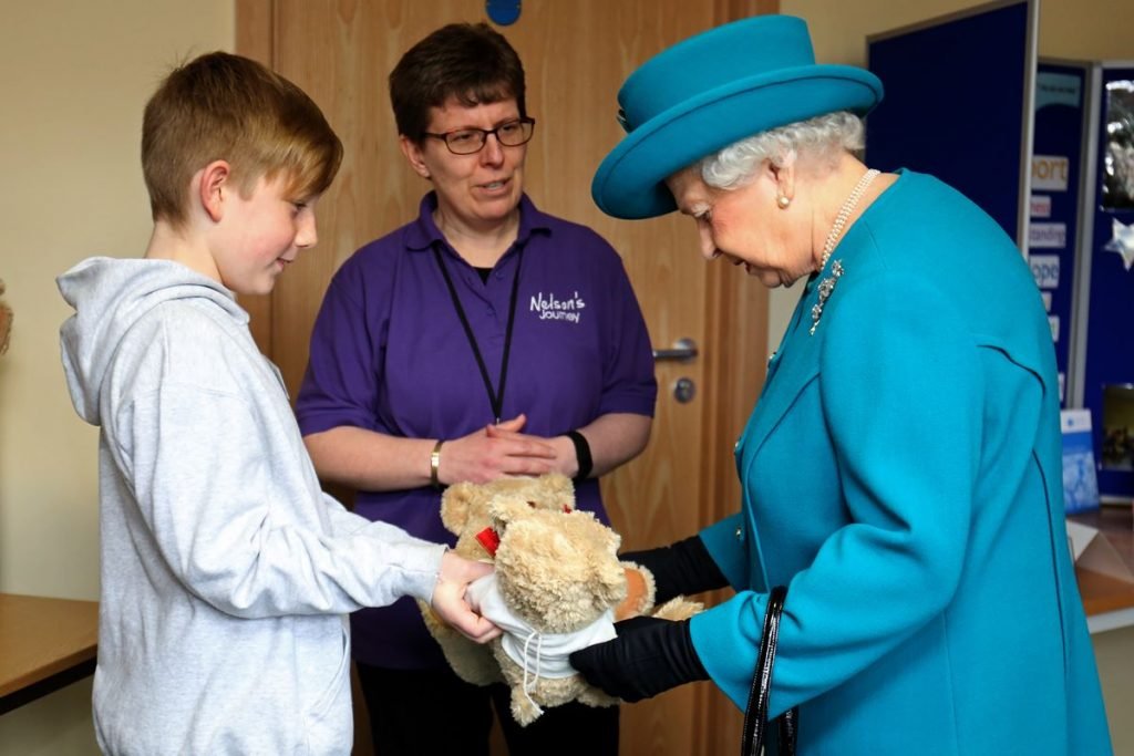 Foto colorida. Rainha Elizabeth recebe de um menino um urso de pelúcia