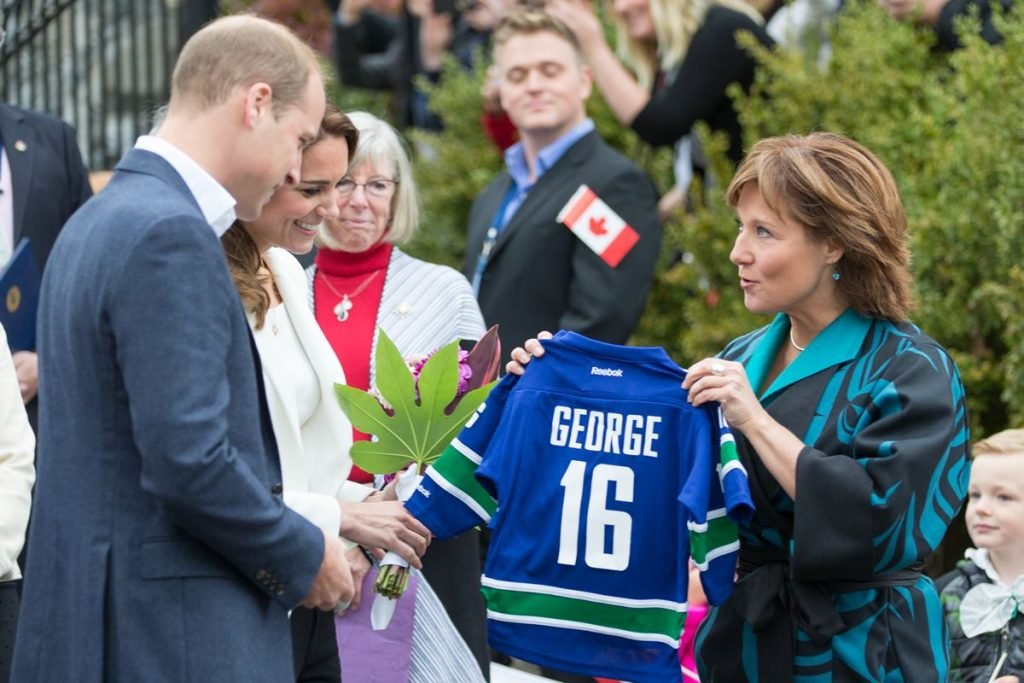 Foto colorida. Príncipe William e Kate Middleton recebem de uma mulher uma blusa com o nome George