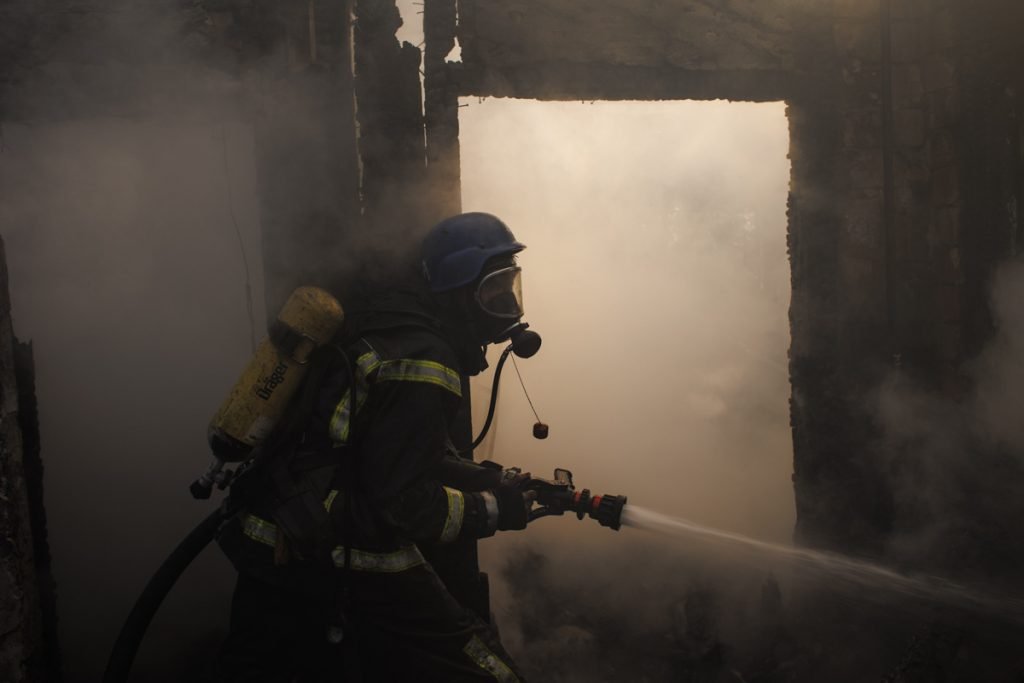 Bombeiros apagam incêndio causado por bombardeio russo na capital ucraniana, Kiev. Na foto um deles apaga fogo com jato de água, uniformizado, dentre muita fumaça - Metrópoles