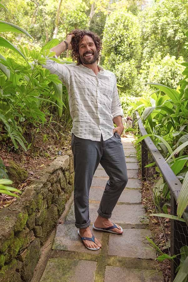 Dudu Azevedo, ator, homem branco de cabelo médio e barba castanha. Ele usa uma calã cinza, camisa branca e chinelos da marca Cartago. Posa para foto em um jardim.