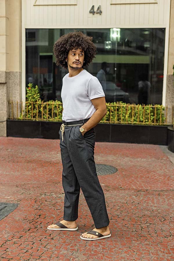 O influenciador Fabianos Gomes, homem negro de cabelo black, posa para foto na faixada de um bloco. Ele usa camiseta branca, calça cinza e chinelos da marca Cartago.