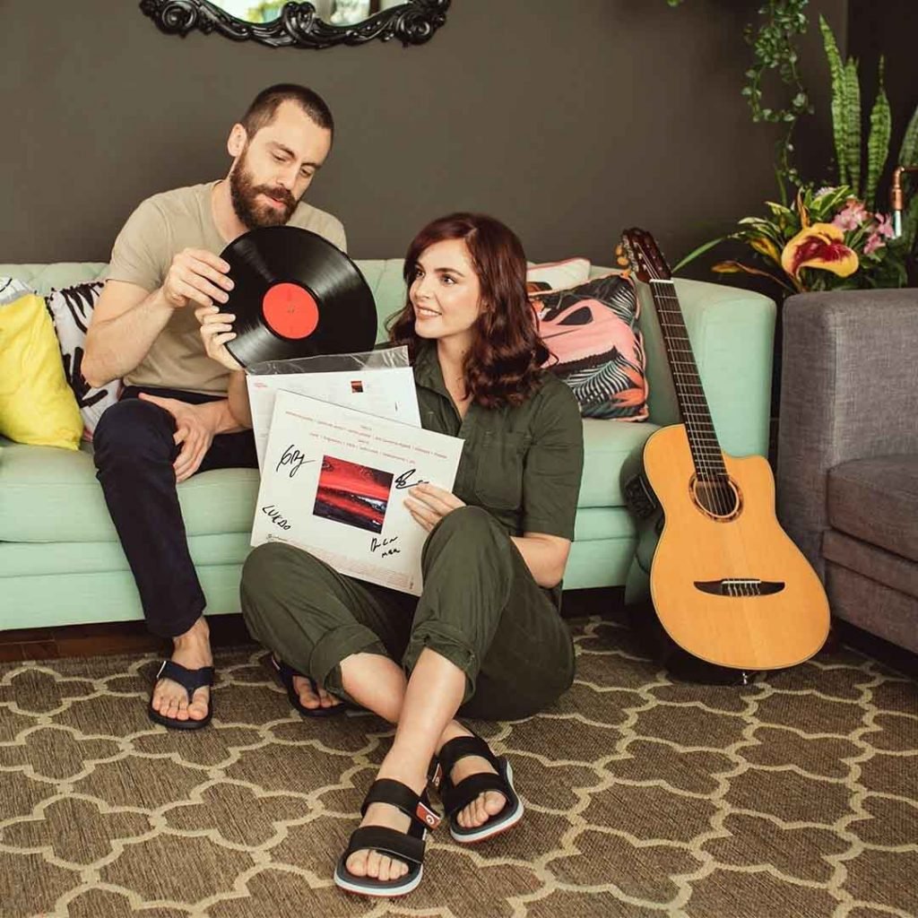 Tomás Bertoni, da banda Scalene, e Titi Muler sentados em um sofã olhando discos de vinil. Ao lado deles é possível ver um violão e outros objetos de decoração