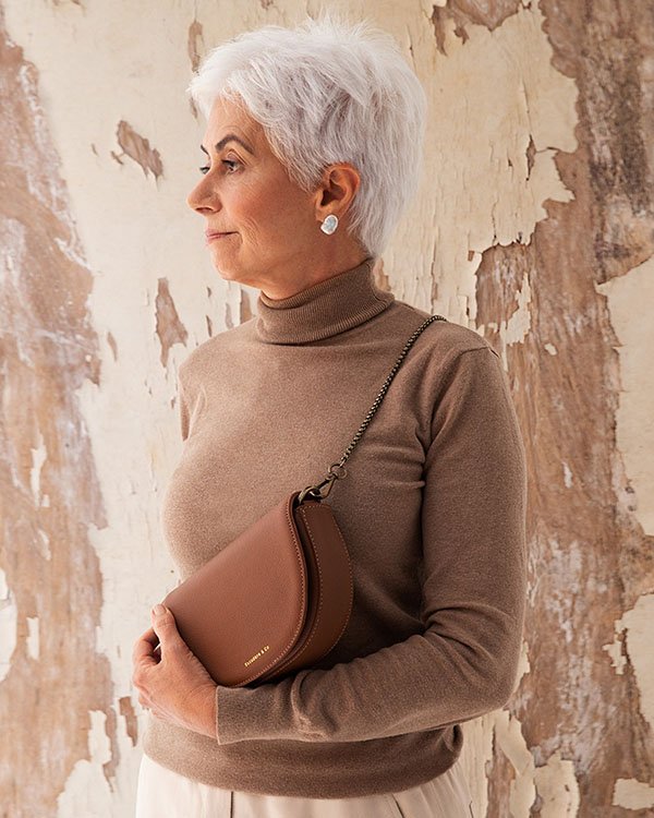 Mulher idosa branca de cabelos curtos grisalhos. Ela usa uma blusa marrom de manga cumprida, na cor marrom, e uma bolsa transpassada de couro da mesma cor. A peça é da marca Escudero & Co.
