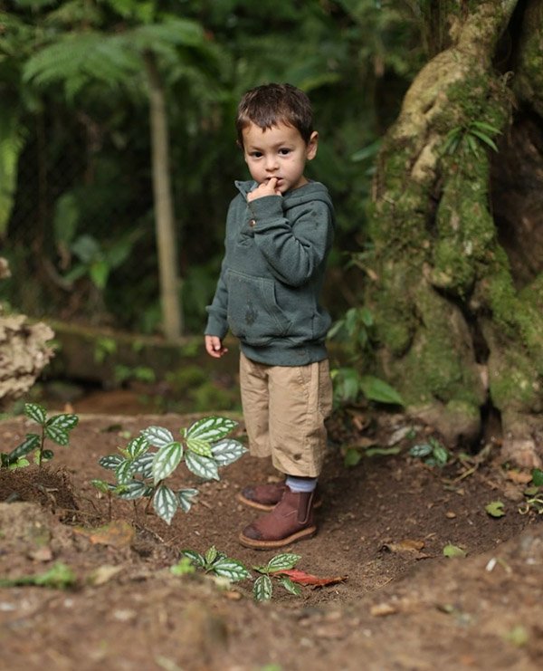 Criança branca brincando na floresta. Ele usa uma calça marrom, casaco cinza e bota da marca João de Barro Botinas.