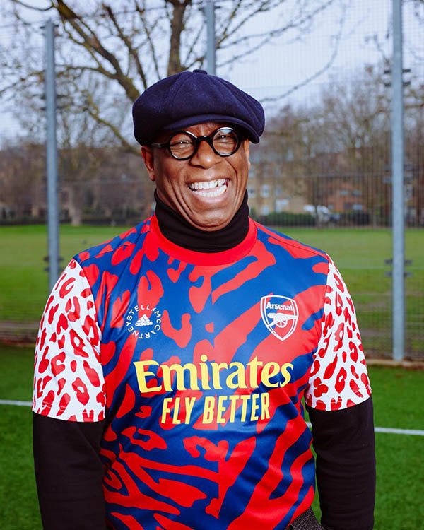 Ian Wright, homem negro careca e ex-jogador do time inglês Arsenal, sorrindo em um campo de futebol. Ele usa óculos de grau, uma boina preta e blusa de manga cumprida preta. Por cima, usa o novo uniforme da equipe feminina do Arsenal, desenhada pela estilista Stella McCartney.