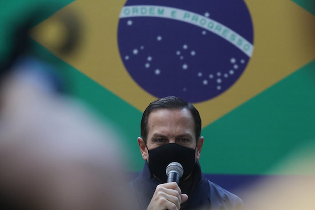 Governador de São Paulo, João Dória discursa de máscara em evento com bandeira do Brasil ao fundo. Ele segura um microfone e usa casasco - Metrópoles