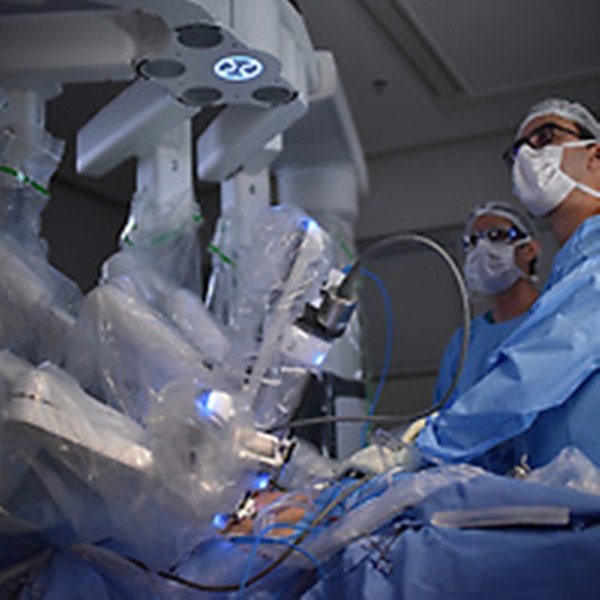 Foto colorida onde um profissional da saúde vestindo uma roupa azul, opera algum maquinário na sala de cirurgia