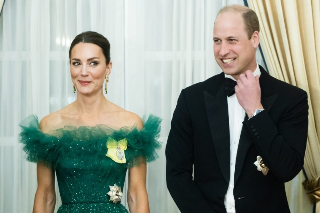 Foto colorida.  Kate Middleton em um vestido verde e o príncipe William em um terno.  Ele pega a gravata