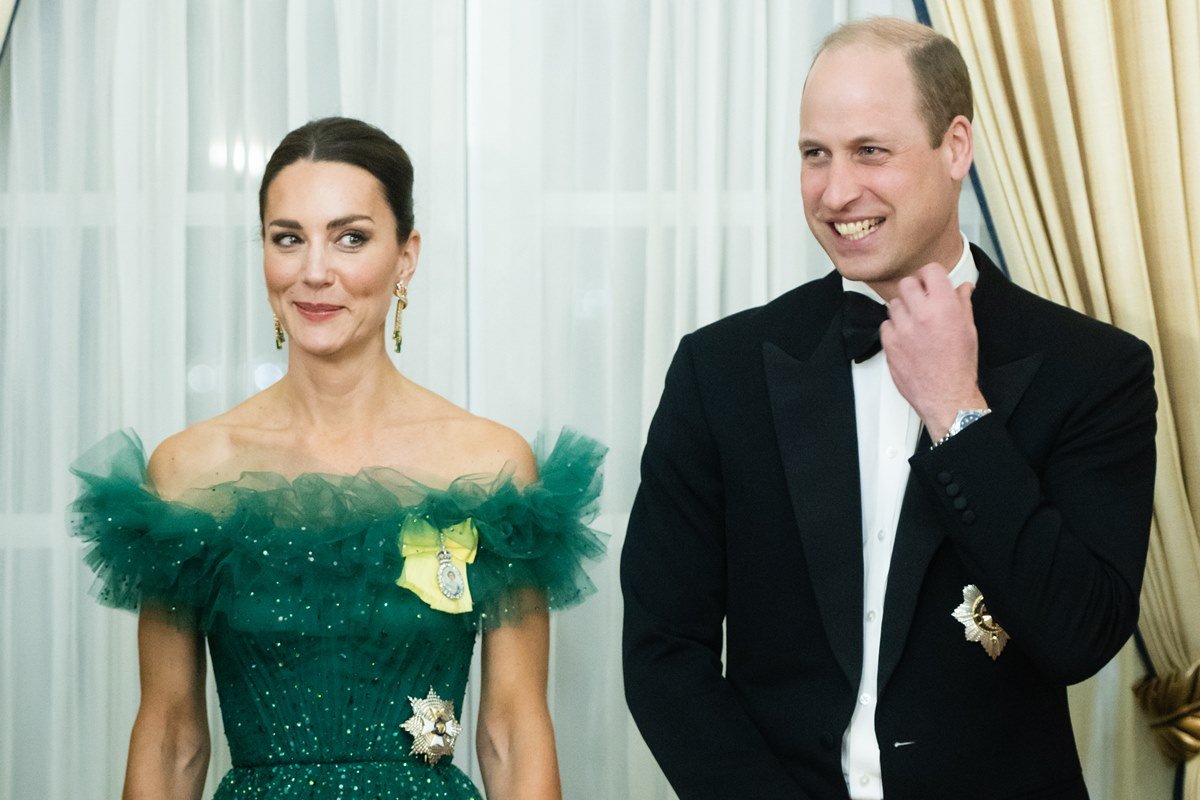 Foto colorida. Kate Middleton de vestido verde e o príncipe William de terno. Ele pega na gravata