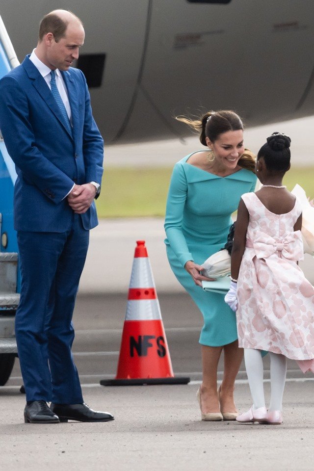 Foto colorida.  Príncipe William e Kate Middleton recebem de uma menina um presente.  William está em pé ao lado