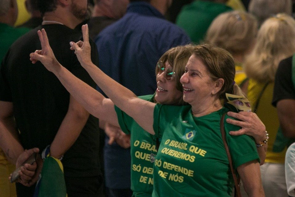 Duas mulheres com camisas verdes e letras em amarelo fazem o sinal do V
