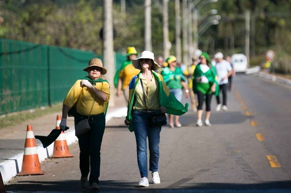 Duas mulheres com camisas amarelas, calça jeans, chapéus e bandeira do Brasil nos ombros caminham por uma pista de asfalto