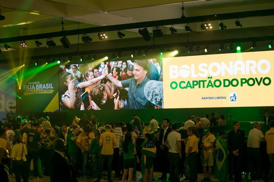 Telão mostra a imagem do presidente Jair Bolsonaro em meio a muita gente.  Ao lado está escrito: 