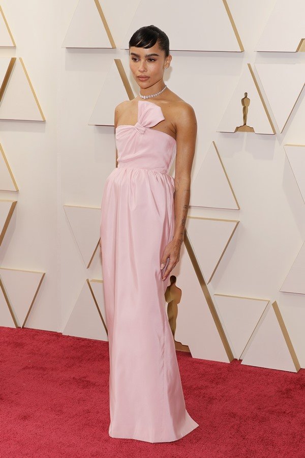 A atriz Zoë Kravitz no tapete vermelho do Oscar 2022. Ela é uma mulher negra, com cabelo presto e uma franja curta, e usa um vestido rosa claro da marca Yves Saint Laurent