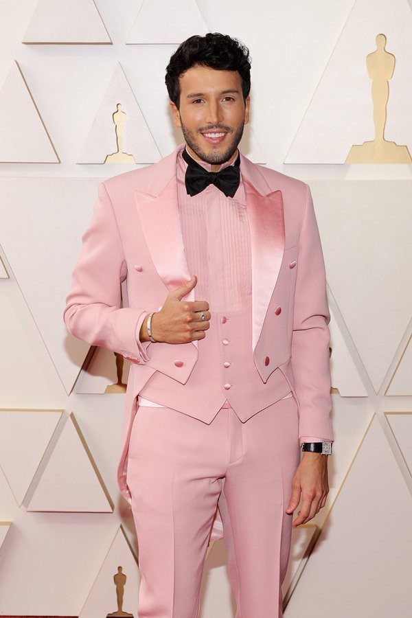 O cantor Sebastián Yatra no tapete vermelho do Oscar 2022. Ele é um homem branco, com cabelo curto e barba preta, e está usando um terno rosa com gravata borboleta preta.