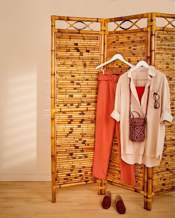 Biombo de madeira com roupas da Amaro pendurada. É possíve ver uma camisa branca de botões no cabide, uma calça coram e uma bolsa com estampa de cobra.