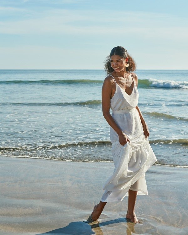 Mulher negra, com cabelo cacheado preso baixo, na praia. Ela usa um vestido branco esvoaçante se alcinha da marca Amaro.