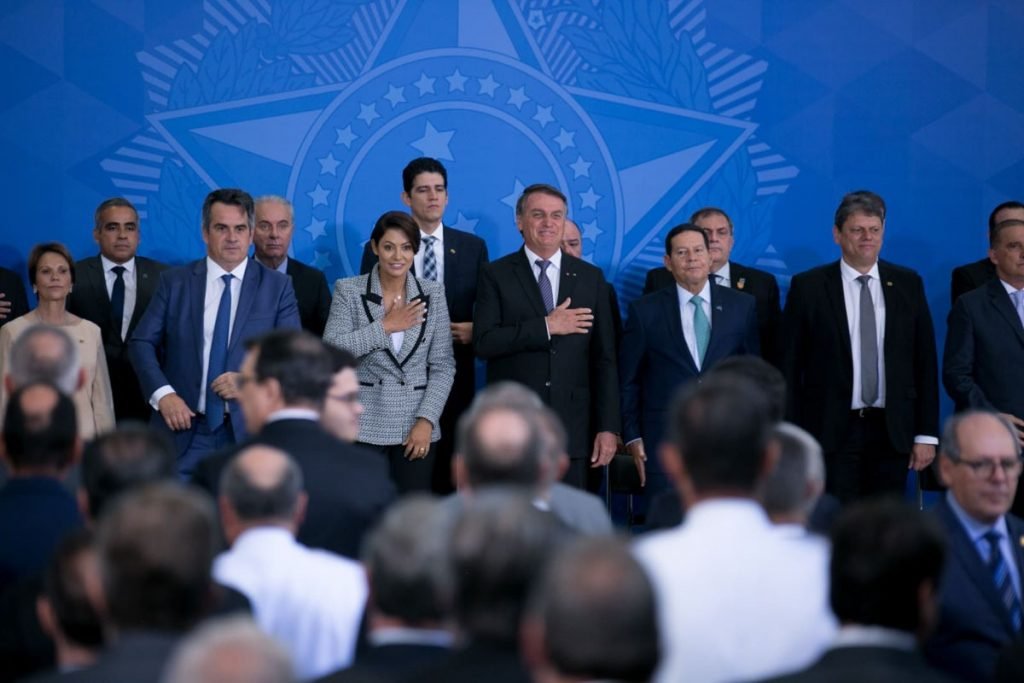 Bolsonaro em cerimônia de despedida aos ministros que serão candidatos nas eleições. Ele leva a mão ao peito ao lado da fileira de ministros e em frente a plateia no Planalto - Metrópoles