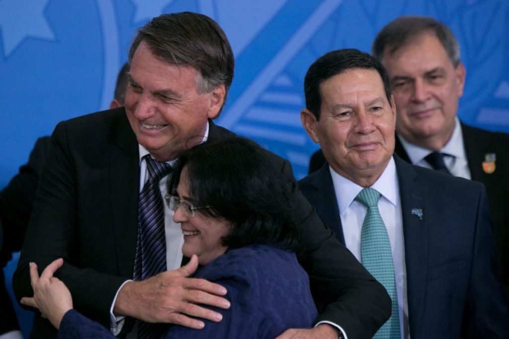 Bolsonaro abraça a agora ex-ministra Damares Alves ao lado do vice-presidente Hamilton Mourão, eme evento de despedida dos ministros no Planalto. O presidente sorri - Metrópoles