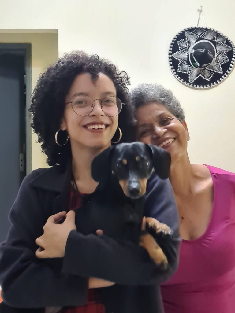 A estudante aprovada em Engenharia Ambiental na UnB, Maria Lívia, ao lado de sua mãe e o cachorrinho da família. Elas sorriem - Metrópoles