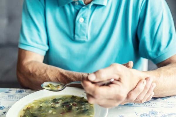 home idoso segura seu próprio braço com a outra mão para tentar comer uma sopa. ele tem Parkinson - Metrópoles